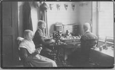 SA1426 - Photo shows three women sewing, a sewing table, and a treadle sewing machine. Identified on the back.